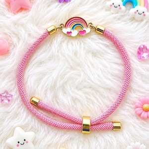 May include: A pink braided cord bracelet featuring a central metal charm of a rainbow with white clouds and two small pink hearts. The bracelet includes gold-toned metal end caps and a sliding gold-toned bead for adjustable sizing and closure.