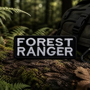 May include: A black rectangular patch with the words "FOREST RANGER" embroidered in white. The patch is displayed on a mossy log with a backpack and carabiner in the background. Ferns and forest foliage surround the patch.