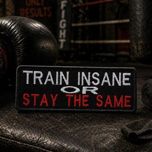 Può includere: Una toppa nera con le parole "TRAIN INSANE OR STAY THE SAME" in lettere bianche e rosse. La toppa è su una superficie strutturata, con guantoni da boxe e una fasciatura arrotolata sullo sfondo.