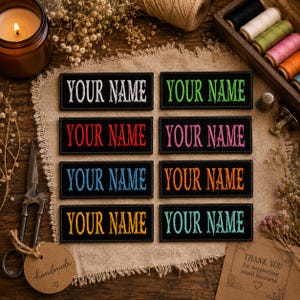 Peut inclure: Huit écussons noirs brodés avec le texte "YOUR NAME" en différentes couleurs : blanc, vert, rouge, rose, bleu, orange, jaune et turquoise. Les écussons sont présentés sur une surface en tissu beige.