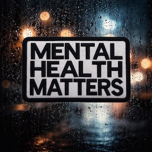 May include: A rectangular patch with the words "MENTAL HEALTH MATTERS" in bold black letters on a white background. The patch has a black border. The background is a blurred image of a rainy window with city lights.