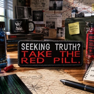 Puede incluir: Una composición con un parche negro con el texto "SEEKING TRUTH? TAKE THE RED PILL", junto a frascos de pastillas rojas y azules. También se ve una taza negra con un conejo, evocando un tema enigmático.