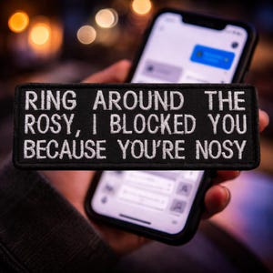 May include: A black rectangular patch with white embroidered text: "RING AROUND THE ROSY, I BLOCKED YOU BECAUSE YOU'RE NOSY." The patch is held in front of a smartphone displaying a messaging app.