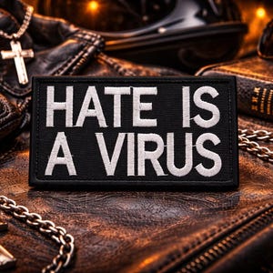 May include: A black patch with the words "HATE IS A VIRUS" in white lettering. The patch is on a leather surface, with a cross necklace, gloves, sunglasses, and a book in the background.
