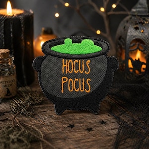 Puede incluir: Parche de caldero negro con contenido burbujeante verde y las palabras "Hocus Pocus" en naranja. El caldero tiene dos asas y tres patas pequeñas. El parche está sobre una superficie de madera con una vela y otras decoraciones de Halloween en el fondo.