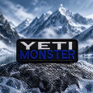 May include: A rectangular black patch with the words "YETI MONSTER" in white and blue lettering. The patch is set against a backdrop of snow-capped mountains and a lake.