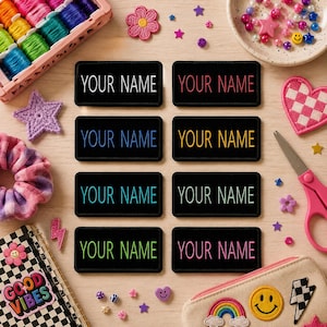 Może przedstawiać: Czarne, prostokątne naszywki z napisem "YOUR NAME" w różnych kolorach. Na zdjęciu znajdują się również materiały do rękodzieła, w tym szpulki nici, notatnik z napisem "GOOD VIBES" i elementy dekoracyjne.