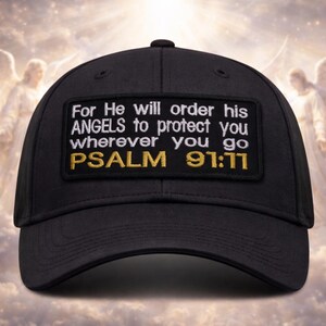 Op de afbeelding: Zwarte baseballcap met een rechthoekige patch met witte tekst die luidt: "For He will order his ANGELS to protect you wherever you go PSALM 91:11" in witte en gouden letters. De achtergrond toont een bewolkte hemel met engelenfiguren.