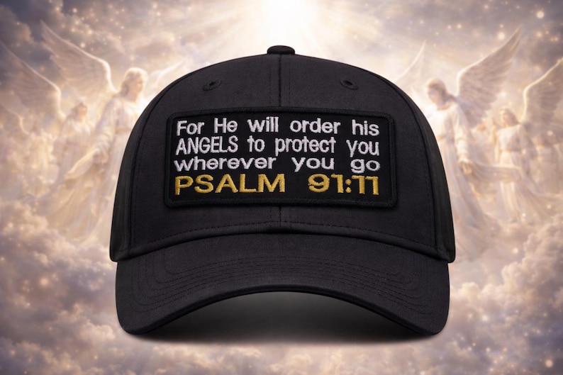 Op de afbeelding: Zwarte baseballcap met een rechthoekige patch met witte tekst die luidt: "For He will order his ANGELS to protect you wherever you go PSALM 91:11" in witte en gouden letters. De achtergrond toont een bewolkte hemel met engelenfiguren.
