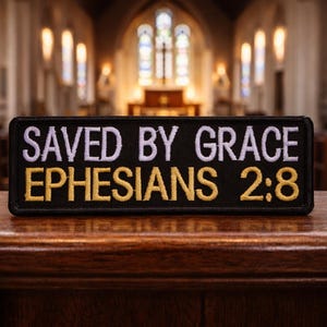 May include: A black rectangular patch with the words "SAVED BY GRACE EPHESIANS 2:8" embroidered in white and gold thread. The patch is displayed on a wooden surface, with a church interior in the background.