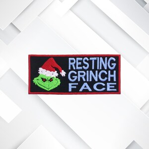 May include: Embroidered patch featuring the Grinch wearing a Santa hat. The patch has a black background with the words "RESTING GRINCH FACE" in light blue. The Grinch is green with a red and white Santa hat.