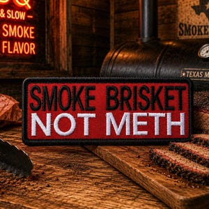 May include: A patch with the text "SMOKE BRISKET NOT METH" in white letters on a red background. A sliced brisket sits on a wooden cutting board. A smoker and a neon sign that says "LOW & SLOW - REAL SMOKE REAL FLAVOR" are in the background.