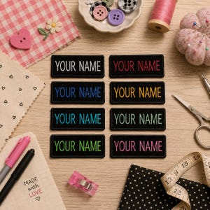 Op de afbeelding: Een verzameling zwarte, geborduurde patches met de tekst "YOUR NAME" in verschillende kleuren. De patches liggen op een houten oppervlak, omringd door naai-attributen zoals knopen, garen en een schaar. Een notitieblok met de tekst "MADE WITH LOVE" is ook zichtbaar.