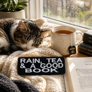 Könnte beinhalten: Eine gemütliche Szene mit einer schlafenden Tabby-Katze auf einer cremefarbenen Decke, einer Tasse Earl Grey Tee und einem Buch. Ein Aufnäher mit den Worten "RAIN, TEA & A GOOD BOOK" ist zu sehen. Die Szene spielt an einem Fenster mit Regen.