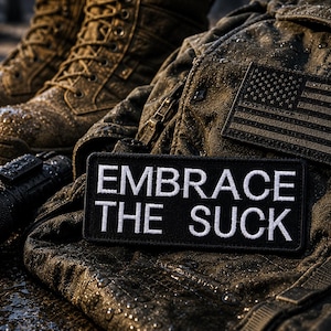 May include: Close-up of military-style gear, including brown combat boots, a tactical jacket, and a black patch with the text "EMBRACE THE SUCK" in white. An American flag patch is also visible.