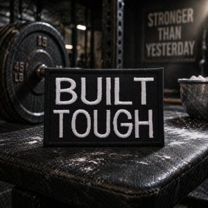 Könnte beinhalten: Schwarzer rechteckiger Aufnäher mit weiß gesticktem Text „BUILT TOUGH“. Der Aufnäher befindet sich auf einer strukturierten Oberfläche, mit einem Gewicht und den Worten „STRONGER THAN YESTERDAY“ im Hintergrund. Das Bild hat ein Fitnessstudio-Thema.