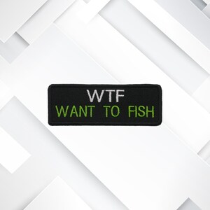 Op de afbeelding: Zwart rechthoekig embleem met de witte tekst "WTF" boven de groene tekst "WANT TO FISH". Deze geborduurde patch is ontworpen om op kleding of uitrusting te worden bevestigd.