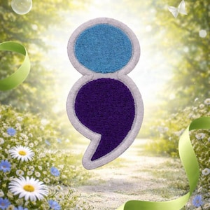 May include: A decorative patch in the shape of a semicolon, with a light blue top and a deep purple bottom. The patch has a white border and a textured, plush appearance. The background features a blurred garden scene with flowers and ribbons.