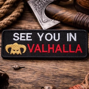 May include: A black embroidered patch with the words "SEE YOU IN VALHALLA" in white and red. The patch features a gold-colored horned skull design. The patch is rectangular and is on a wooden surface.