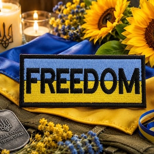 Puede incluir: Un parche rectangular con la palabra "FREEDOM" en letras negras sobre un fondo azul y amarillo. La imagen también presenta girasoles, velas y placas de identificación militares, con una cinta azul y amarilla.