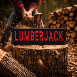 Puede incluir: Un parche rectangular negro con la palabra "LUMBERJACK" bordada en rojo. El parche se muestra sobre un tocón de árbol, con una persona cortando leña en el fondo. La escena está ambientada al aire libre.