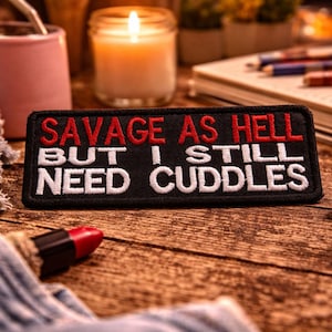 May include: A black rectangular patch with the words "SAVAGE AS HELL BUT I STILL NEED CUDDLES" embroidered in red and white. The patch is on a wooden surface, with a lit candle, a pink cup, and a pair of headphones in the background.