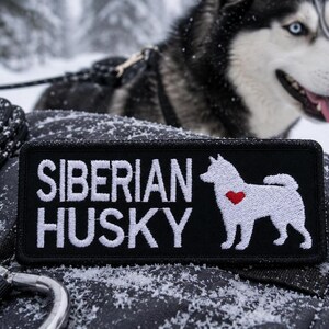 以下が含まれることがあります： 白い文字で「SIBERIAN HUSKY」と刺繍された黒いパッチ。赤いハートが付いた犬の白いシルエットが特徴です。背景には、白と黒のハスキー犬がいます。