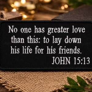 May include: Black rectangular patch with white embroidered text: "No one has greater love than this: to lay down his life for his friends. JOHN 15:13." The patch is on a burlap surface, with a wooden cross and a Bible in the background.