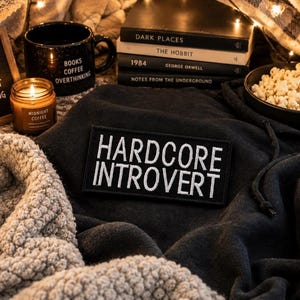 May include: A cozy scene featuring a black mug with "Books Coffee Overthinking" text, a lit candle, and a patch that reads "Hardcore Introvert." Books, popcorn, and a sign saying "Do Not Disturb Introvert Recharging" complete the relaxing aesthetic.