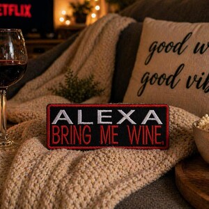 May include: A cozy scene with a glass of red wine, a lit candle, and a patch that reads "ALEXA BRING ME WINE." A pillow with "good wine good vibes" is in the background. Popcorn and a lit candle are also visible.