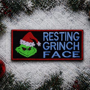 May include: A rectangular patch with a black background and a red border. The patch features a green Grinch face wearing a red Santa hat with white trim. The words "RESTING GRINCH FACE" are embroidered in blue.