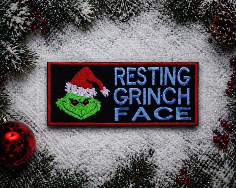 May include: A rectangular patch with a black background and a red border. The patch features a green Grinch face wearing a red Santa hat with white trim. The words "RESTING GRINCH FACE" are embroidered in blue.