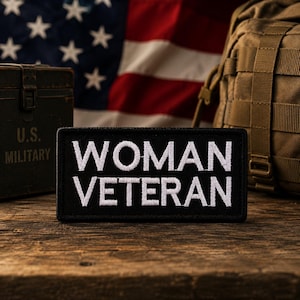 以下が含まれることがあります： 白いブロック体で「WOMAN VETERAN」と書かれた黒いパッチ。パッチは木製の表面にあり、背景にはアメリカ国旗と軍事装備があります。