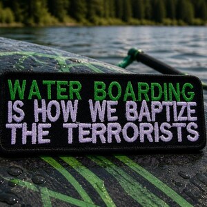 May include: A black rectangular patch with green and white embroidered text that reads "WATER BOARDING IS HOW WE BAPTIZE THE TERRORISTS." The patch is on a dark surface with green and black patterns, with a body of water in the background.