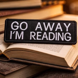 以下が含まれることがあります： 「GO AWAY I'M READING」と白い刺繍が施された黒いパッチが、開いた本の上に置かれています。本は他の本の上にあり、積み重ねられています。画像は暖かく、魅力的なトーンです。
