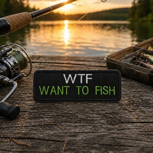 May include: A black embroidered patch with the text "WTF WANT TO FISH" in white and green. The patch is on a wooden surface, with fishing gear and a lake in the background. A fishing rod and reel are visible.
