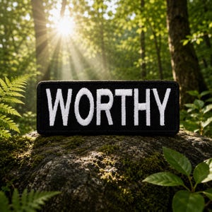May include: A black rectangular patch with the word "WORTHY" embroidered in white. The patch is set against a natural backdrop of a moss-covered rock and lush green foliage, with sunlight streaming through the trees.