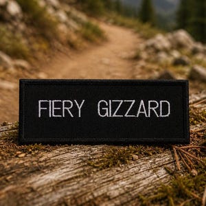 May include: A black rectangular patch with the embroidered text "FIERY GIZZARD" in white. The patch is displayed on a weathered wooden surface with a blurred natural background.