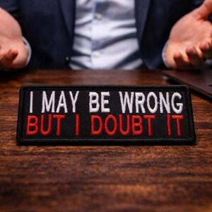 May include: Black rectangular patch with the embroidered text "I MAY BE WRONG BUT I DOUBT IT" in white and red. The patch is on a wooden surface. A person's hands are visible in the background.