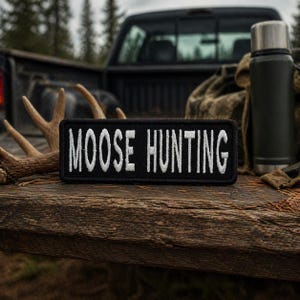 May include: A black rectangular patch with the words "MOOSE HUNTING" embroidered in white. The patch is on a wooden surface, with a truck bed and antlers in the background. A green thermos is also visible.