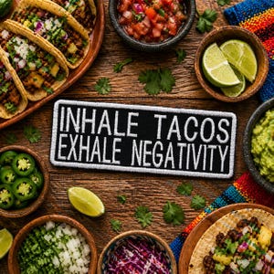 May include: A wooden table is laden with a variety of Mexican food items. Tacos, salsa, guacamole, lime wedges, and chopped vegetables surround a black patch with the text "INHALE TACOS EXHALE NEGATIVITY" in white.