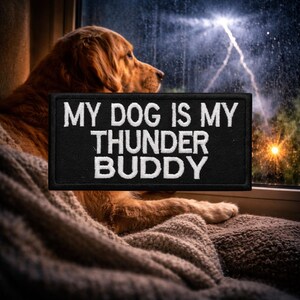 May include: A golden retriever dog rests on a cozy blanket near a window, with a black patch that reads "MY DOG IS MY THUNDER BUDDY" in white lettering. The background shows a stormy sky with lightning.