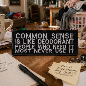 以下が含まれることがあります： 「I survive on coffee and sarcasm」と書かれた白いセラミックマグが、黒いパッチの隣に置かれています。パッチには「Common sense is like deodorant, people who need it most never use it」と書かれています。To-doリストのメモ帳と付箋も表示されています。