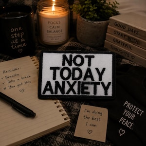 May include: A black and white patch with the words "NOT TODAY ANXIETY" is surrounded by a candle, a notebook, and a small plant. A black mug reads "one step at a time." A small card says "I'm doing the best I can."