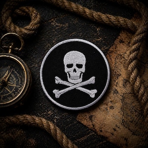 May include: Round black patch with a white skull and crossbones design, bordered by white stitching. The patch is set against a dark, textured wooden surface, with a compass and rope in the background. A classic pirate symbol.