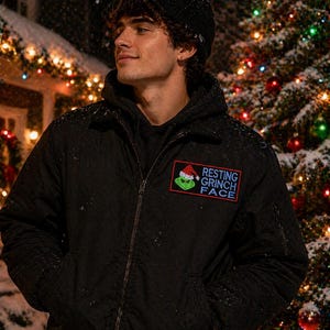May include: Black bomber jacket with a "Resting Grinch Face" patch. The patch features the Grinch wearing a Santa hat. The jacket has a zipper closure and two front pockets. The background includes a Christmas tree with colorful lights and falling snow.