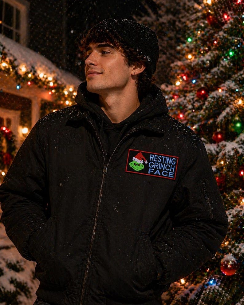 May include: Black bomber jacket with a "Resting Grinch Face" patch. The patch features the Grinch wearing a Santa hat. The jacket has a zipper closure and two front pockets. The background includes a Christmas tree with colorful lights and falling snow.