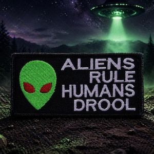 May include: Black rectangular patch with the text "ALIENS RULE HUMANS DROOL" in white. The patch features a green alien head with red eyes. A UFO hovers in the background, with a green beam of light.