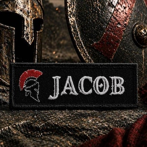 May include: Black rectangular patch embroidered with the name "JACOB" in white, stylized lettering. A red and white Spartan helmet graphic is on the left. A weathered helmet and shield are in the background.