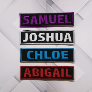 Puede incluir: Cuatro etiquetas bordadas con bordes negros. Los nombres son Samuel en morado, Joshua en blanco, Chloe en azul y Abigail en rojo.
